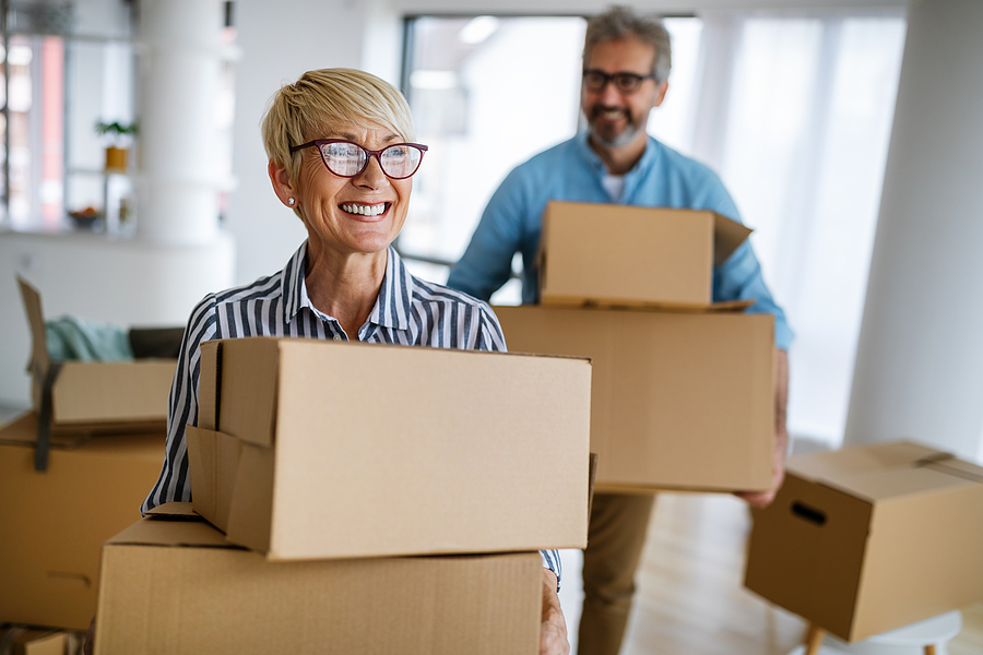 A senior couple smiles while carrying moving boxes.